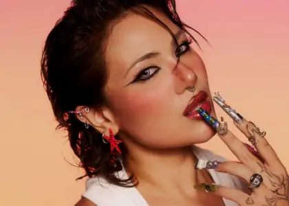 SILVY with bold eyeliner and shiny lips poses thoughtfully, displaying intricate, multicolored nail art and star-shaped earrings, against a warm gradient background.