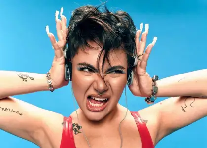 SILVY with short hair and tattoos, wearing headphones against a bright blue background. They look animated and expressive, with open mouth and raised hands.