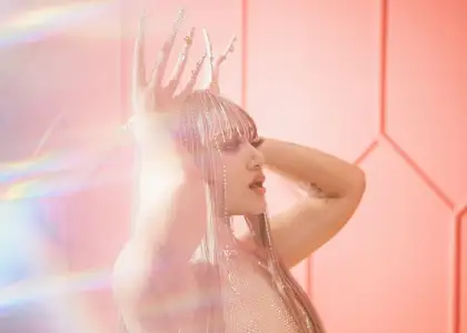 SILVY with long, silvery hair and rhinestone-adorned nails poses confidently against a pink geometric background, conveying glamour and elegance.