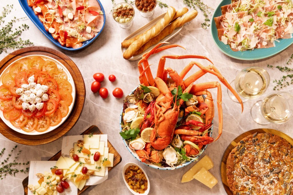 A table set with seafood, cheese, breadsticks, sliced tomatoes, salad, prosciutto, wine, and dessert, arranged attractively for a festive meal.