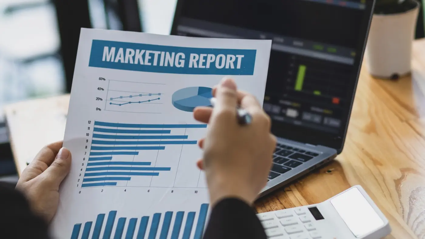 A person holds a paper titled “Marketing Report” with charts and graphs, sitting at a desk with a laptop, calculator, and pen.