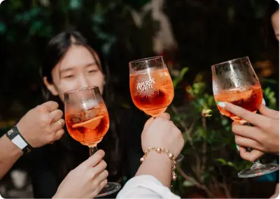 Four people clink glasses of an orange cocktail outdoors. A woman in the background smiles, and greenery surrounds the group, creating a relaxed, festive atmosphere.