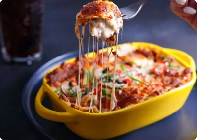 A close-up of a cheesy baked pasta dish in a yellow casserole dish, with a fork lifting a gooey, melted cheese-filled serving. The dish is topped with herbs and vegetables.