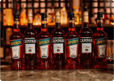 Six bottles of red liqueur—three Aperol and three Campari—are lined up on a bar counter with a blurred background of shelves and warm lighting.