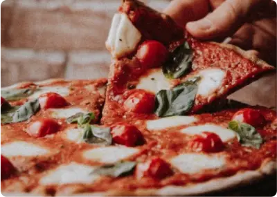 A hand lifts a slice of pizza topped with tomato sauce, cherry tomatoes, mozzarella cheese, and fresh basil leaves. The crust is thin and slightly browned.