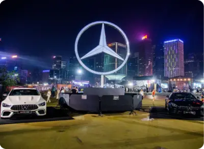 Two Mercedes-Benz cars parked on either side of a large illuminated Mercedes-Benz logo, with a city skyline and skyscrapers in the background at night.