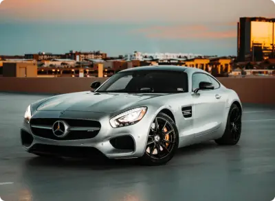 A silver Mercedes-Benz sports car is parked on a rooftop with city buildings and a golden sunset reflecting in the background.