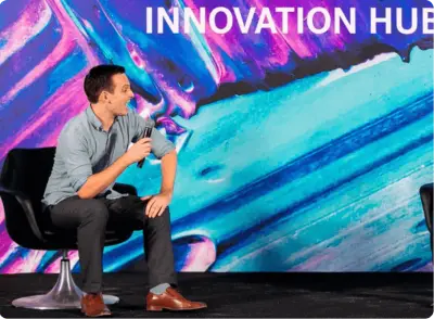 A man sits in a black chair holding a microphone, speaking on stage with a colorful “INNOVATION HUB” sign in the background.