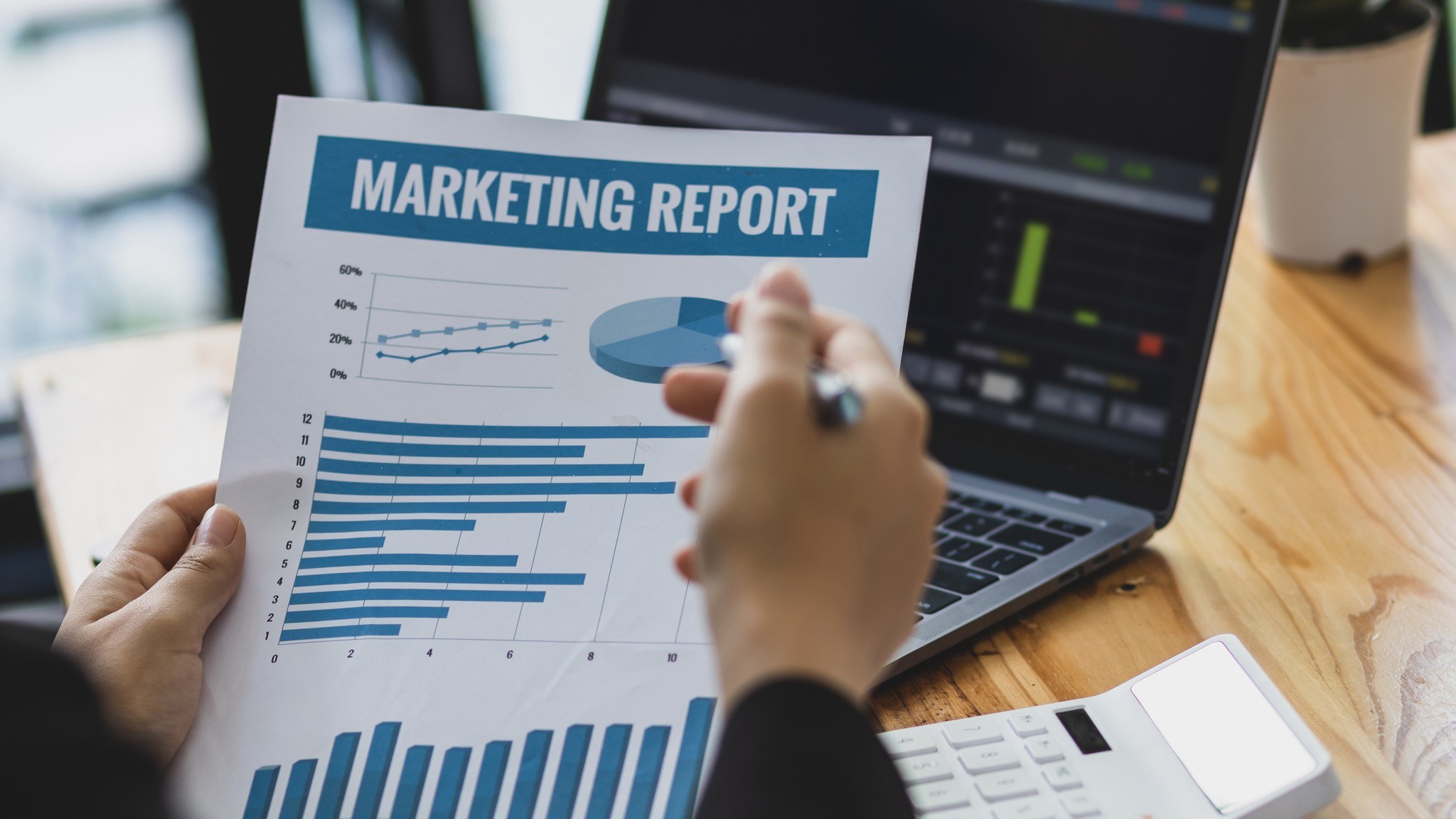 A person holds a paper titled “Marketing Report” with charts and graphs, sitting at a desk with a laptop, calculator, and pen.