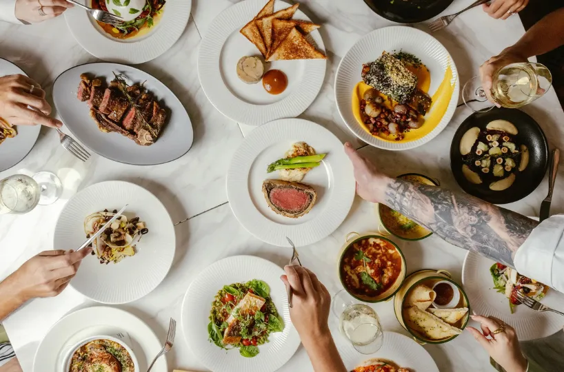 A group of people sharing an elegant meal, featuring steak, toast, salad, roasted vegetables, pasta, and wine, all arranged on a white tablecloth. Multiple hands reach for or serve the dishes.
