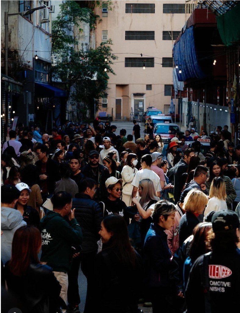 A crowded city street filled with people, some talking and some walking. Tall buildings line the street, and sunlight filters through, casting shadows on the crowd.