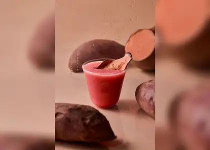 A clear plastic cup filled with pink smoothie, garnished with a sweet potato chip, surrounded by whole and sliced sweet potatoes on a reflective, light-brown surface, with condensation on a blurred backdrop.