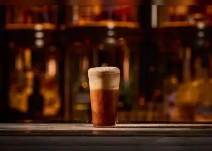 A tall glass of a dark, layered drink with a thick, frothy, tan-colored top, sitting on a bar with a blurred, warm-toned bar interior in the background.