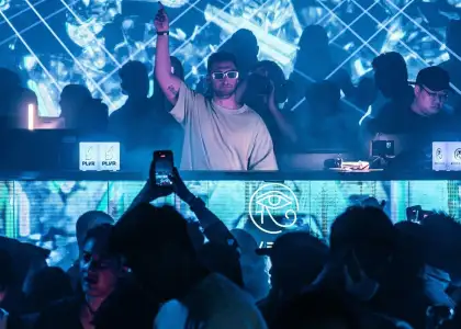 A man stands confidently in front of a lively crowd at a nightclub, illuminated by colorful lights.