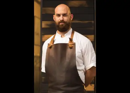 A bearded man in an apron stands in front of a textured wall, smiling and looking towards the camera.