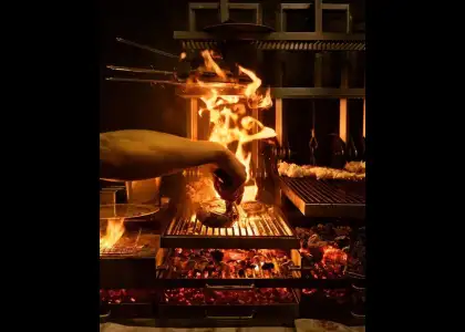 A person grilling food over an open flame, with smoke rising and vibrant flames visible around the cooking surface.