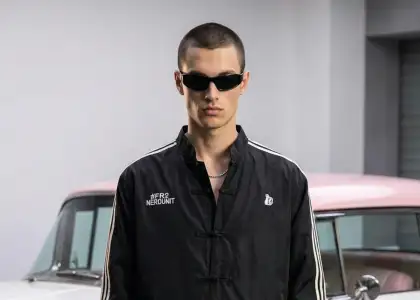 A person in black sunglasses and a black jacket with white stripes stands confidently in front of a vintage car