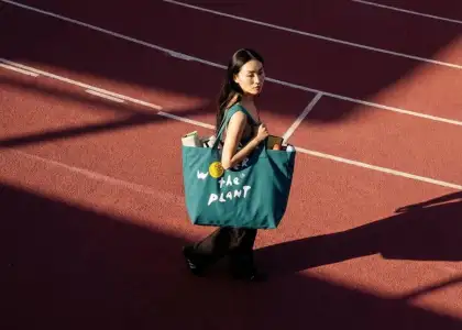 A woman walks on a sunlit athletic track carrying a large teal bag that reads "WATER THE PLANT."