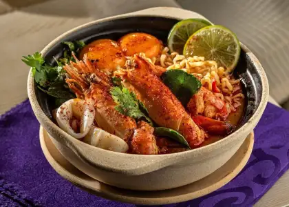 A vibrant bowl of spicy seafood ramen featuring prawns, calamari, boiled eggs, lime slices, and fresh herbs