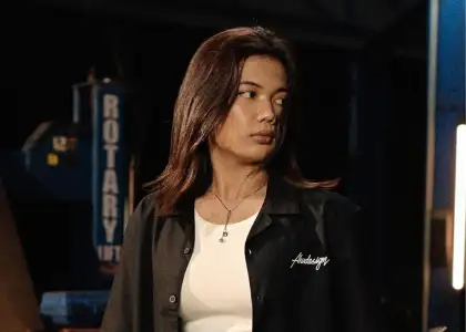 A person with long hair, wearing a black jacket and white shirt, gazes to the side in a dimly lit industrial setting