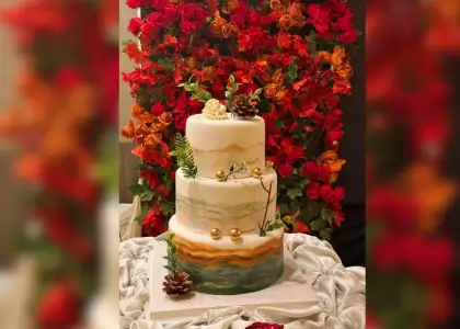 Three-tiered cake with earthy tones, pine cones, and gold accents, set against a vibrant red and orange floral backdrop