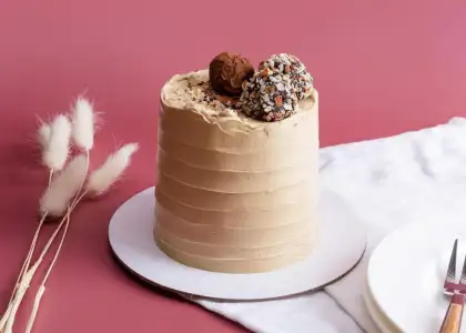Tall cake on a white plate with smooth, light brown frosting, topped with truffles