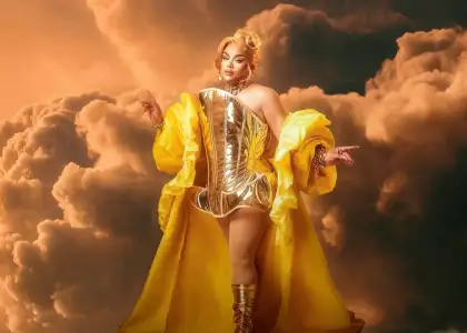 Tiny Deluxe wears metallic gold corset, yellow flowing cape, and matching boots