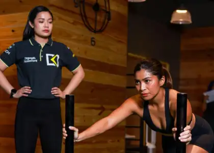 Two women are in a gym with a wooden wall and clock. One is pushing a sled, appearing focused, while the other watches attentively