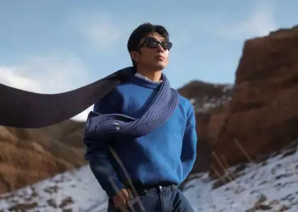 A man in sunglasses and a blue sweater with a scarf poses confidently in a snowy, rocky landscape