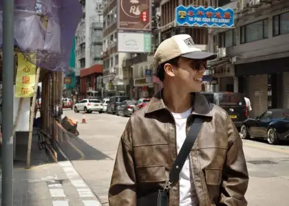 A person in a brown leather jacket and cap smiles while standing on a sunny street