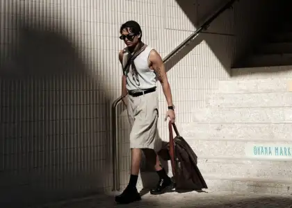 A man in a sleeveless shirt and knee-length shorts walks down sunlit stairs, carrying a large bag