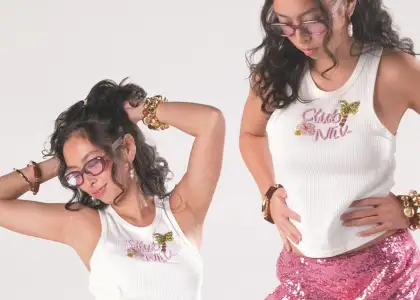 A woman wears white tank tops with floral and butterfly designs, pink sunglasses, and bold gold jewelry