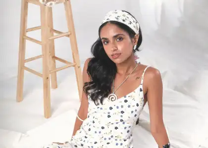 Woman in a floral dress and headband sits on a white fabric backdrop, beside a wooden stool