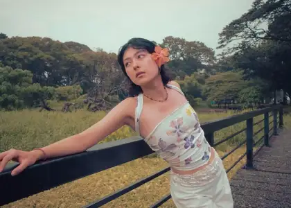 A woman leans on a black railing in a park, looking contemplative. She wears a floral top and a flower in her hair.