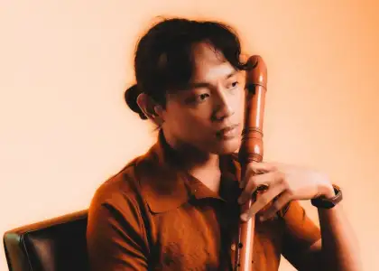 Halina in a rust-colored shirt sits pensively holding a wooden flute against a warm, orange-toned background