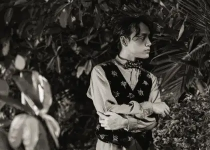 A person stands thoughtfully against a backdrop of lush foliage, wearing a patterned vest and bow tie