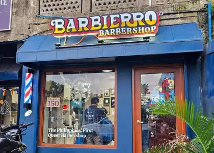 Exterior of a blue barbershop with "Barbierro" sign and rainbow flag. The window displays "The Philippines' First Queer Barbershop," creating an inclusive and welcoming vibe.