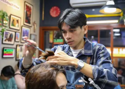 A focused hairstylist in a plaid shirt trims a customer's hair in a cozy salon, with framed art and a red wall clock in the background.
