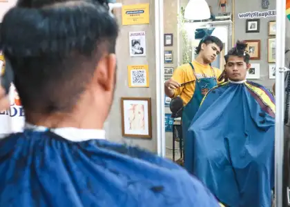 A barber in a yellow shirt and apron cuts a customer's hair in front of a mirror. The customer, wearing a blue cape, looks directly at the reflection. The scene is calm and focused, with framed pictures on the wall adding a personal touch.