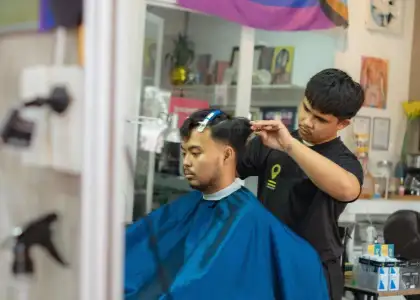 A barber focuses intently while trimming a man's hair in a salon. The man wears a blue cape and looks relaxed. The scene is calm and professional.