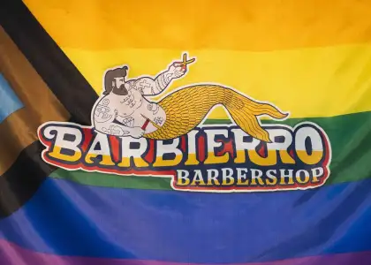Colorful LGBTQ+ flag background with "Barbierro Barbershop" logo featuring a tattooed merman holding scissors. Bold, inclusive, playful theme.