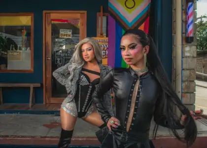 Two drag performers pose confidently outside a barbershop with a Pride flag. One wears silver and black, the other in sleek black, exuding empowerment.