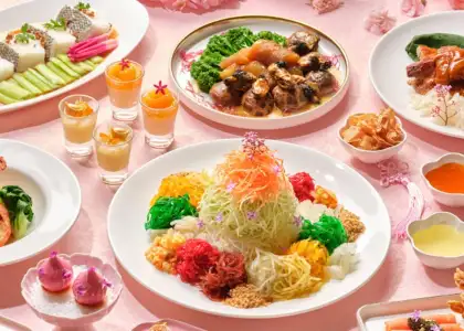 A spread of Chinese New Year dishes, including a colorful salad in the center, various small appetizers and desserts, and steaming cups of tea, all arranged on a deep red tablecloth