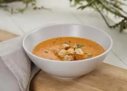 Bowl of creamy tomato soup topped with croutons and parsley, placed on a wooden board with a gray napkin
