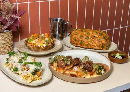A table with gourmet dishes includes a pastry-topped loaf, meat-topped ravioli, broccoli Alfredo, and a floral centerpiece
