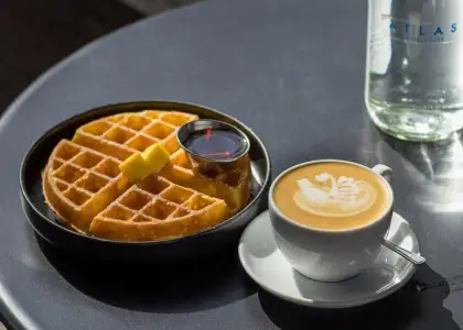 A black plate with a golden waffle topped with butter, a small syrup cup, and a cappuccino with latte art on a dark table
